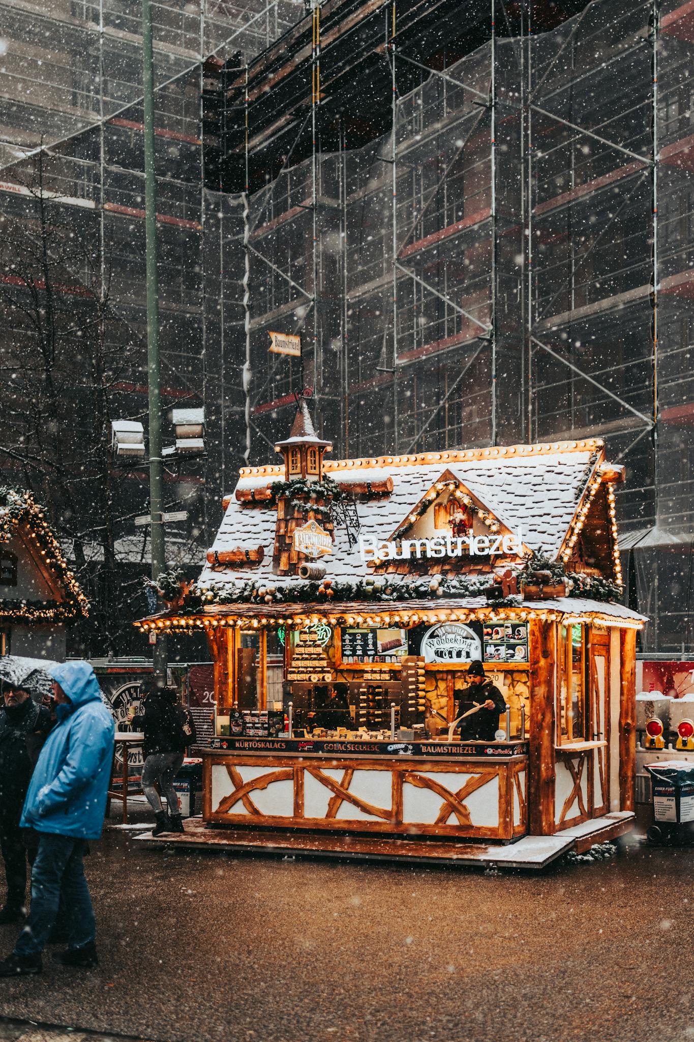 Illuminated Christmas market stall during snowfall in the city, showcasing festive decorations and lights.